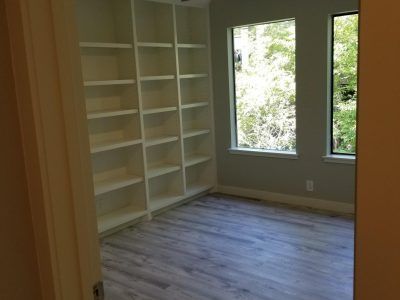 A room with built-in white shelves and two windows overlooking greenery; gray wood-look flooring.