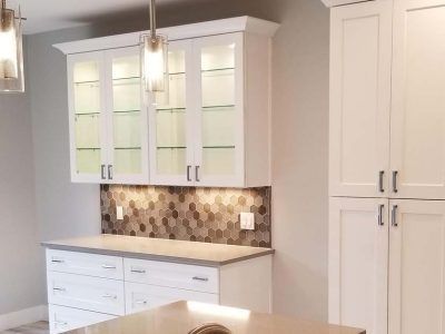 White kitchen cabinets with glass doors, illuminated interior, gray backsplash, and countertop.