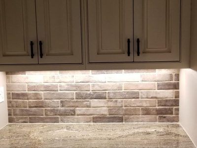 Brick backsplash beneath white kitchen cabinets with black handles; countertop in front.