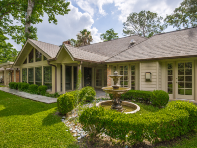 Lush green yard with a fountain in front of a tan brick and glass-windowed house.