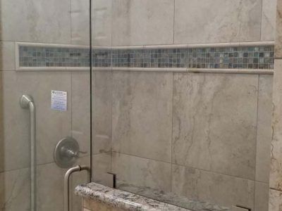 Shower stall with light-colored tile, glass door, and mosaic tile accent. A grab bar and faucet are visible.