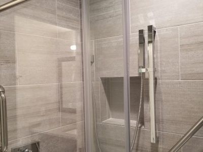 Shower interior with glass door, recessed shelf, and silver fixtures on gray tiled walls.