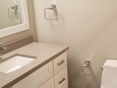 Bathroom with white vanity, sink, mirror, and towel rings. Beige countertop and off-white walls.