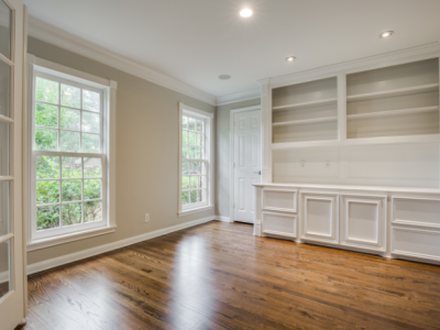 Empty room with hardwood floors, built-in shelving, windows, and white trim.
