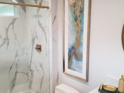 Bathroom with marble shower, gold fixtures, art, and white walls.