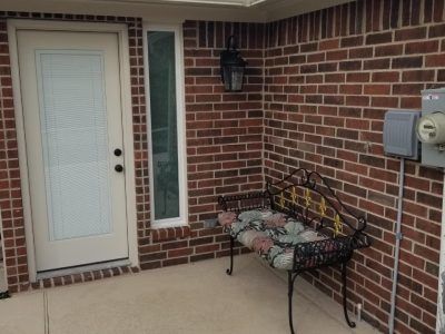 Brick-walled patio with white door, narrow window, black bench with floral cushion, and electrical box.