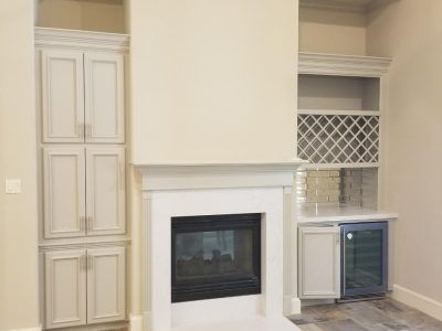Fireplace flanked by built-in cabinets in a neutral-colored room. One cabinet has a wine rack and a mini-fridge.