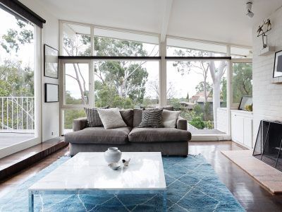 Living room with large windows overlooking trees. A gray sofa, blue rug, and white marble coffee table.