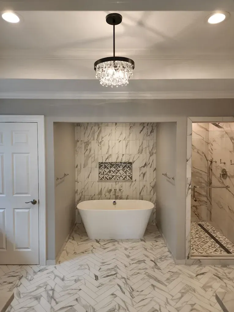 Bathroom with freestanding tub set in a marble-tiled alcove, crystal chandelier, and herringbone floor.