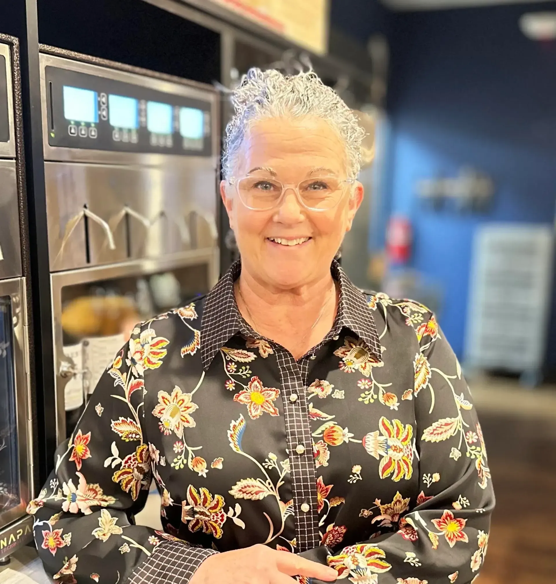 Woman with silver hair smiles, wearing glasses and floral shirt, stands near a beverage machine.