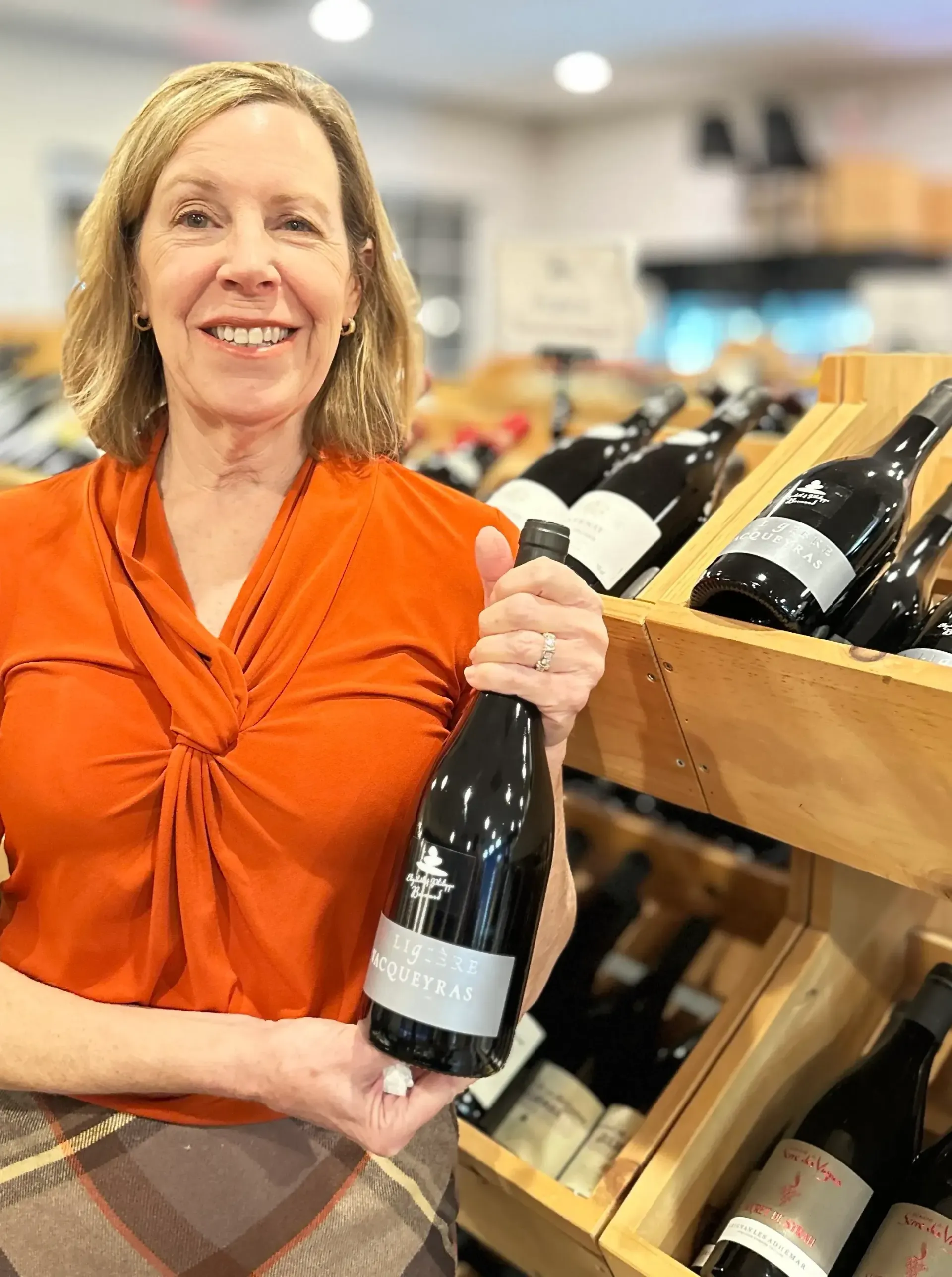 Woman in orange shirt holds a wine bottle in a store, smiling near shelves of wine.