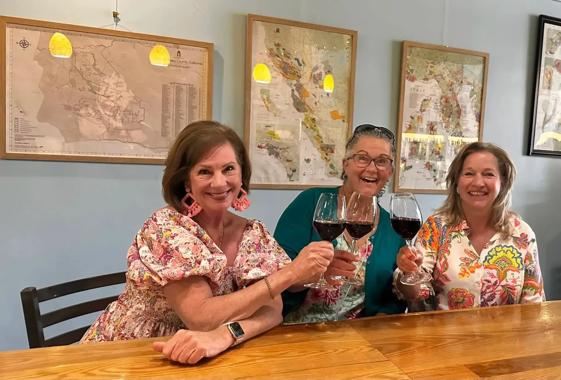 Three women toasting with red wine at a bar. Maps hang on the wall behind them.