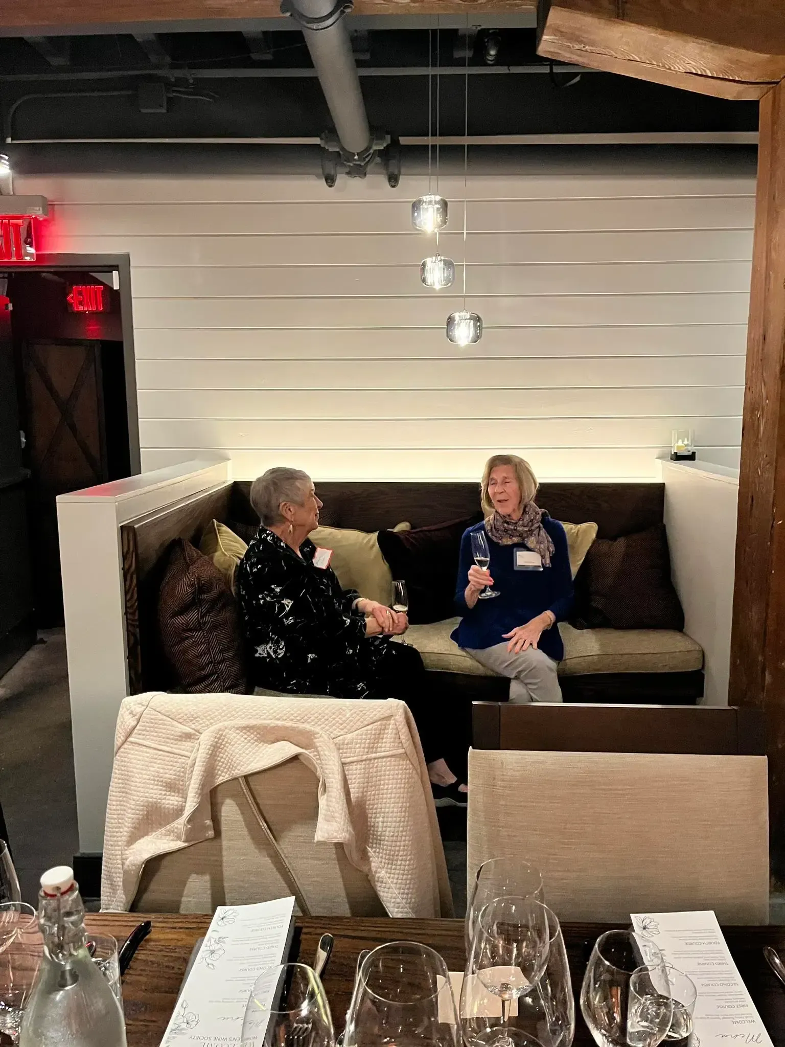 Two women seated in a booth, one speaking, possibly an interview. Restaurant setting, neutral tones.