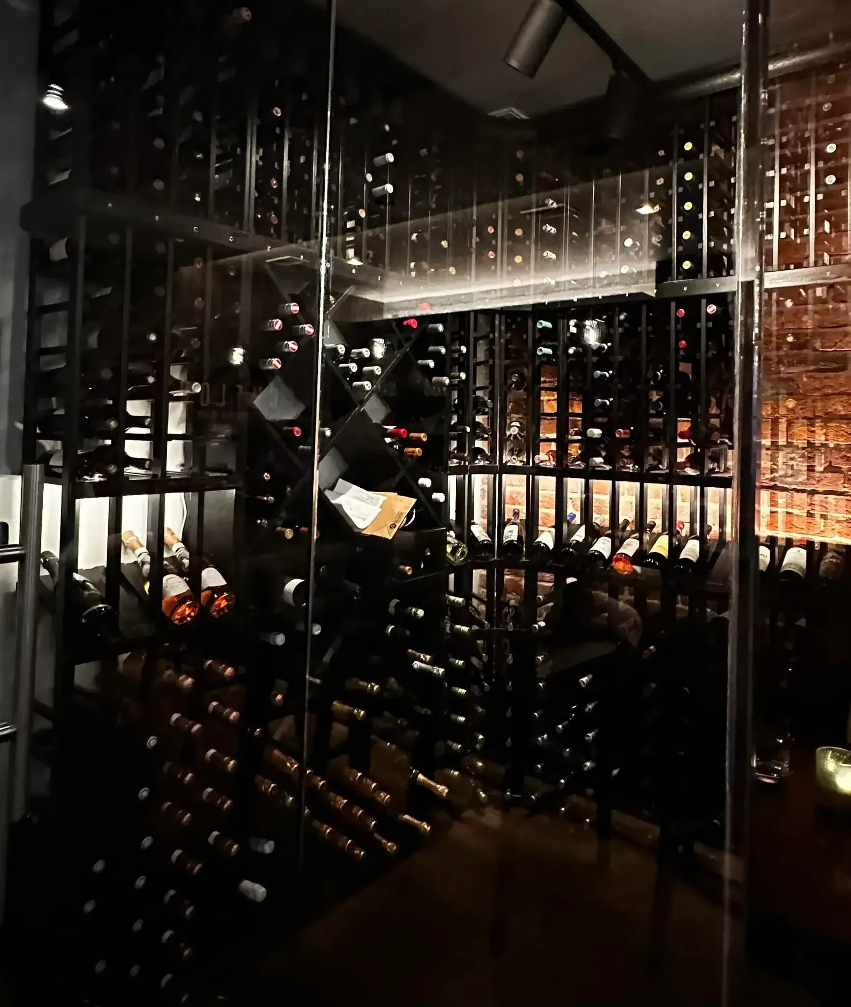 Man in black shirt holding wine glass, speaking at restaurant table. Yellow wall, painting, diners.