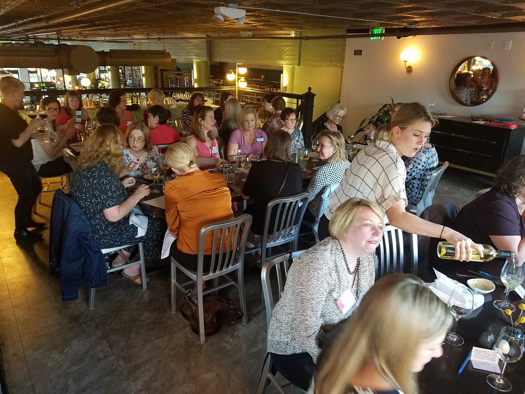People at tables in a restaurant, a woman pouring wine, other women smiling and chatting.