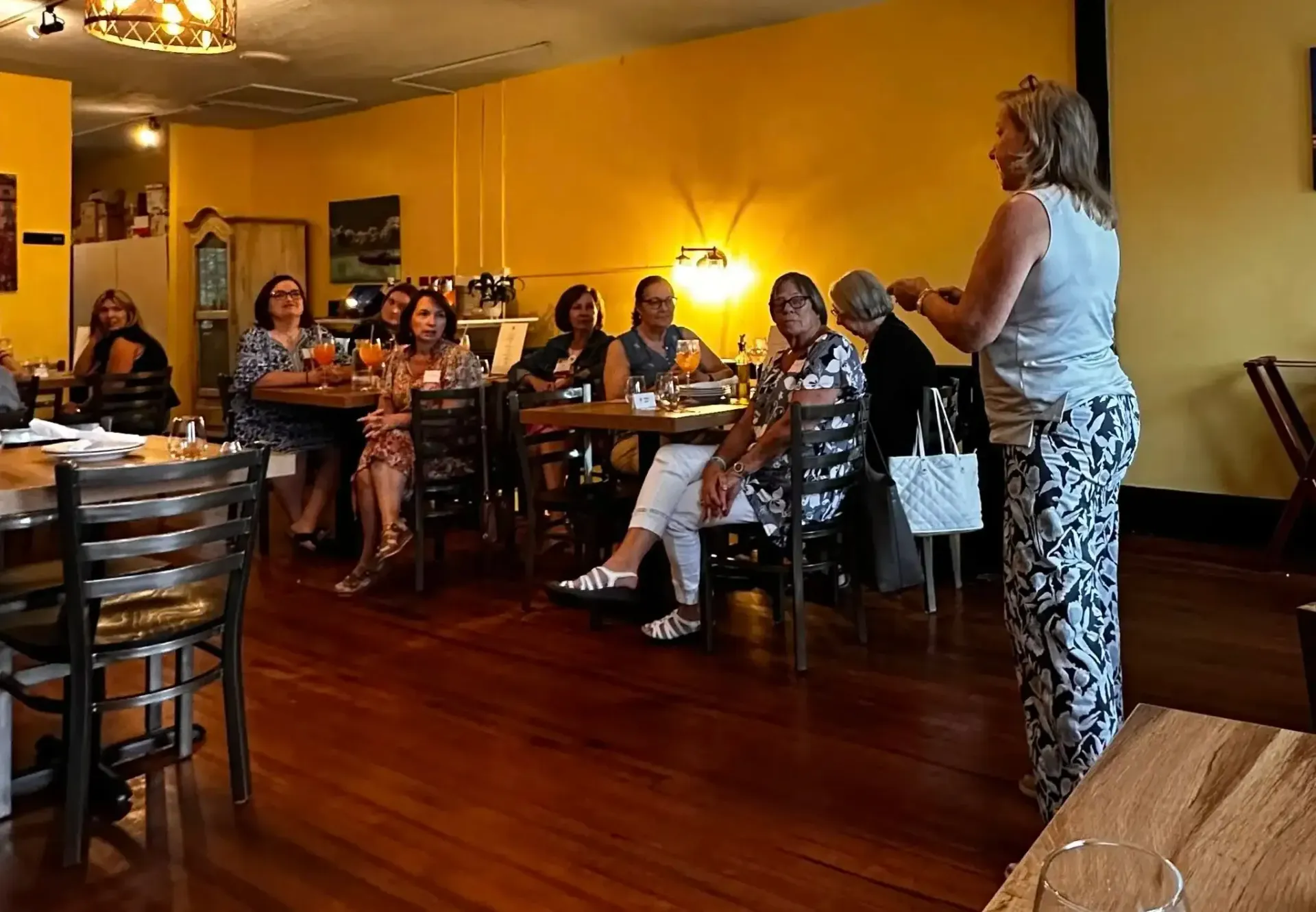 Woman giving a presentation to a group seated at tables in a yellow-walled restaurant.