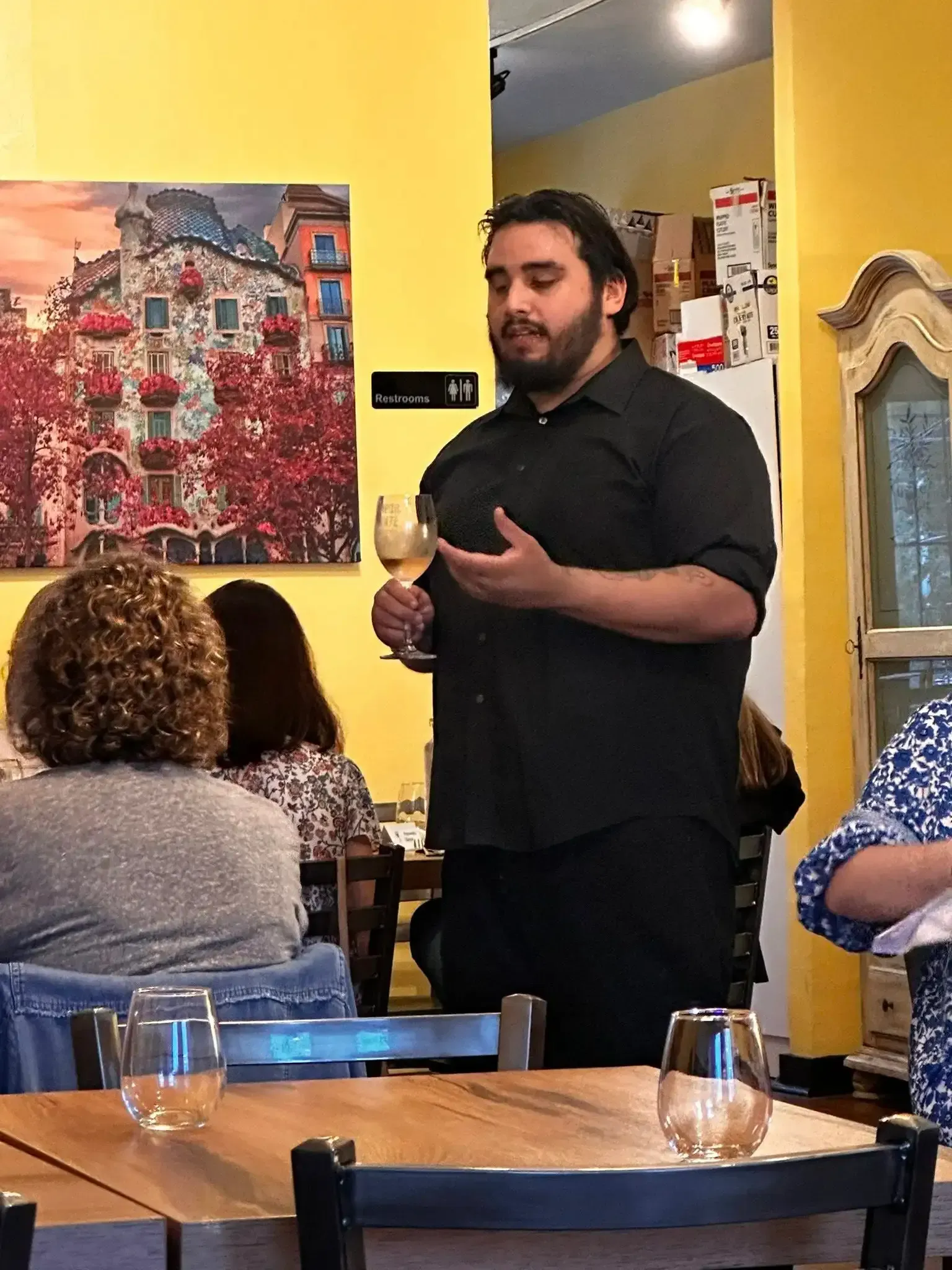 Man in black shirt holding wine glass, speaking at restaurant table. Yellow wall, painting, diners.