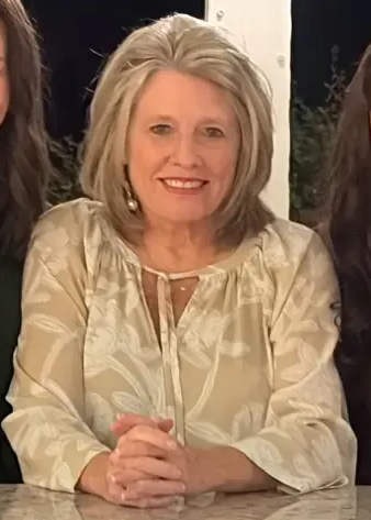 Woman with graying hair smiles, hands clasped. She wears a light-colored top, indoors at a table.
