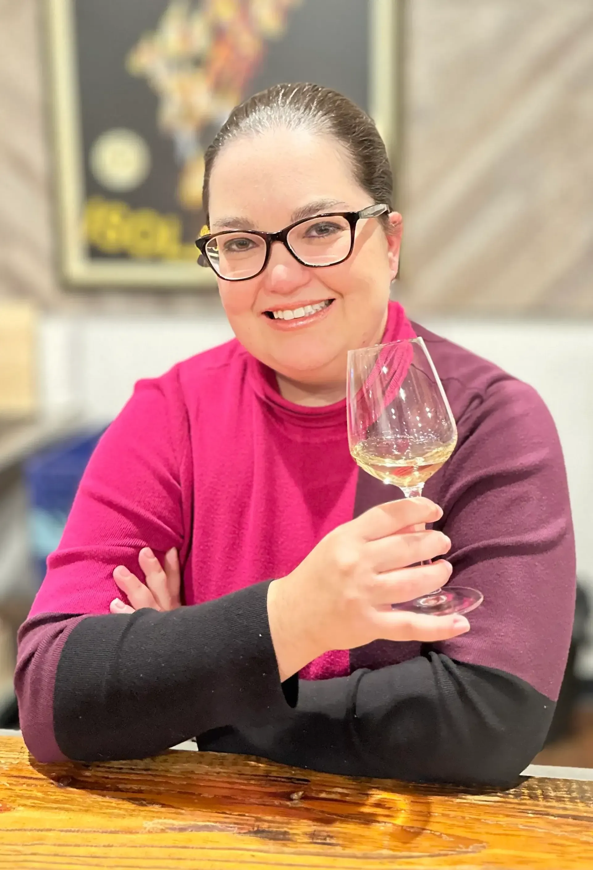 Woman with glasses holding wine glass, smiling. Wearing pink and black sweater, indoors.