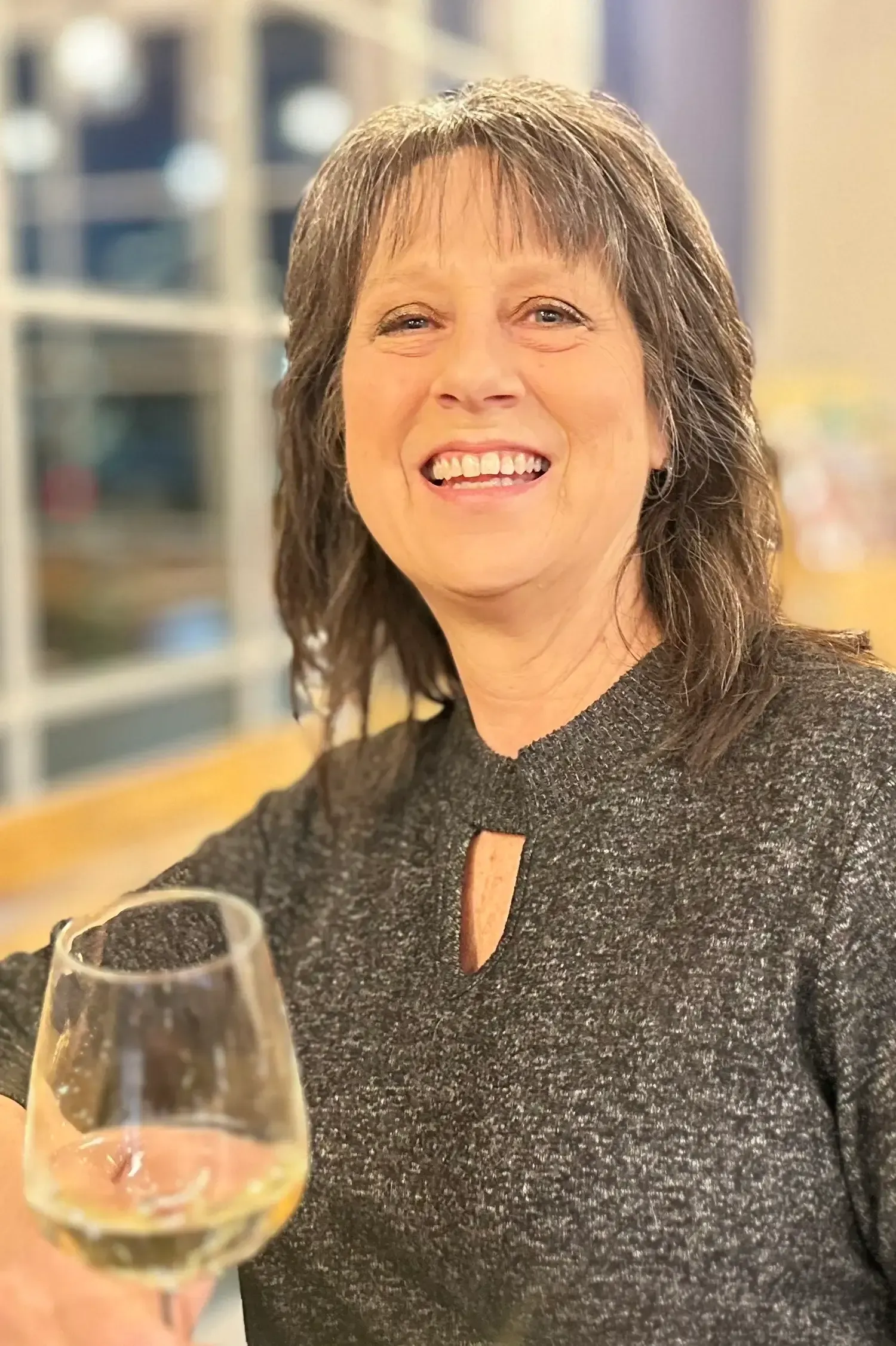 Woman smiles, holding a wine glass. She wears a gray sweater with a keyhole neckline, in a brightly lit setting.