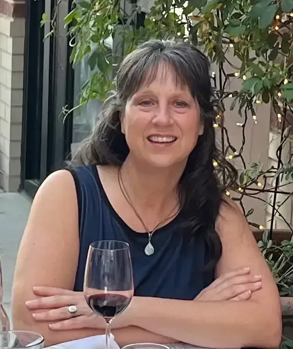 Woman smiles, holding a wine glass. She wears a gray sweater with a keyhole neckline, in a brightly lit setting.