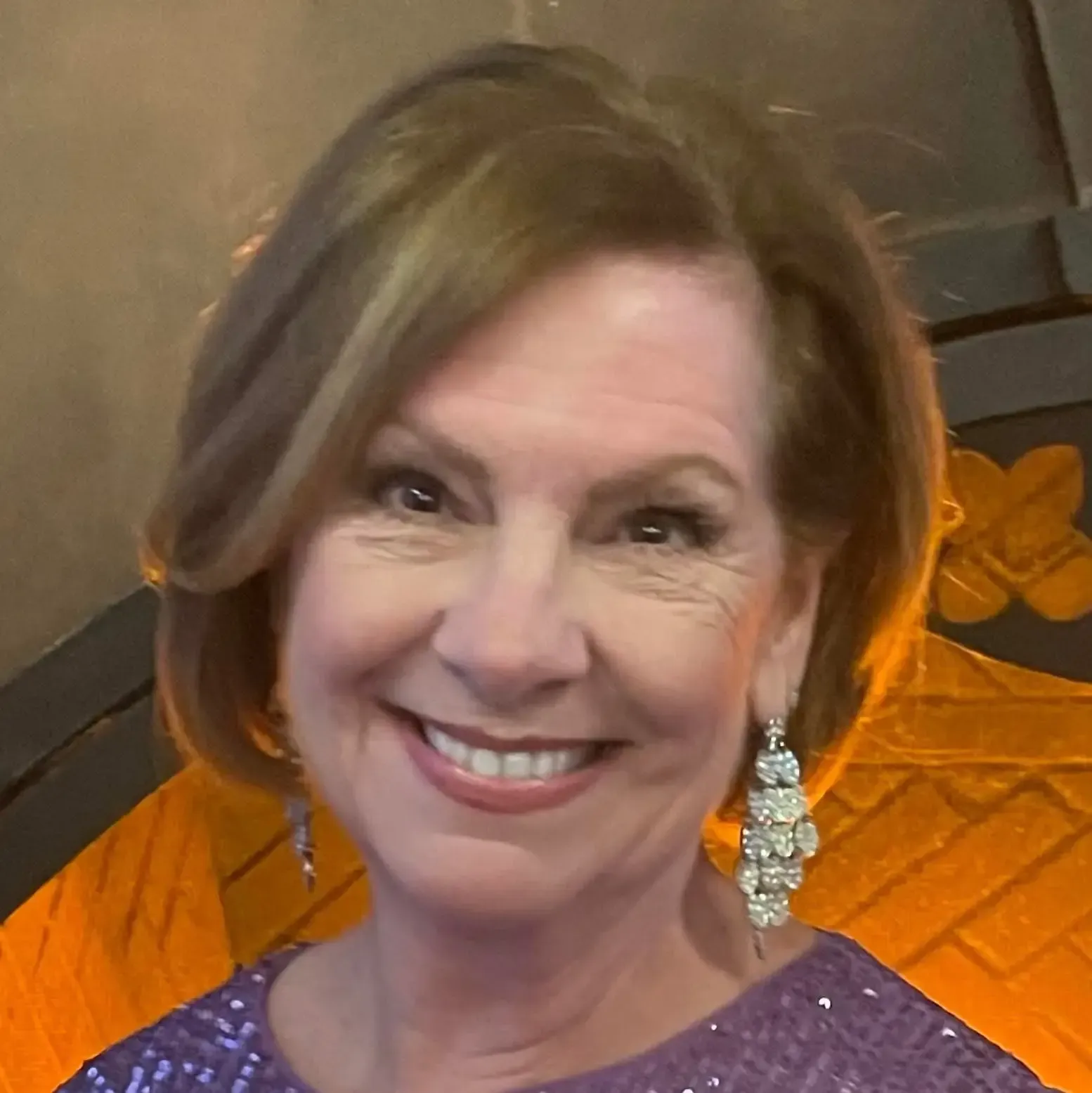Woman with short brown hair smiles, wearing sparkling earrings and purple sequined top.