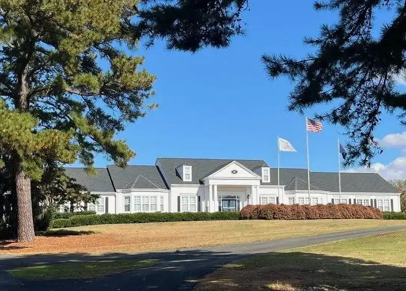White building with flags against a blue sky, framed by green trees and bushes.