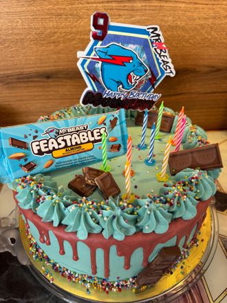 Een blauwe verjaardagstaart met glazuur, versierd met snoep, lolly's, chocoladerepen en een Beastable-figuurtje bovenop een tafel.