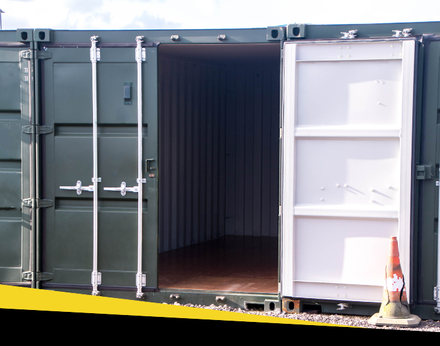 An open, dark green shipping container with a white inner door panel and an orange safety cone on the ground.