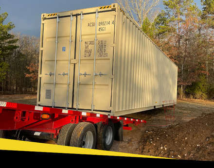 A tan shipping container is being transported on a red flatbed trailer parked on a dirt lot near trees.