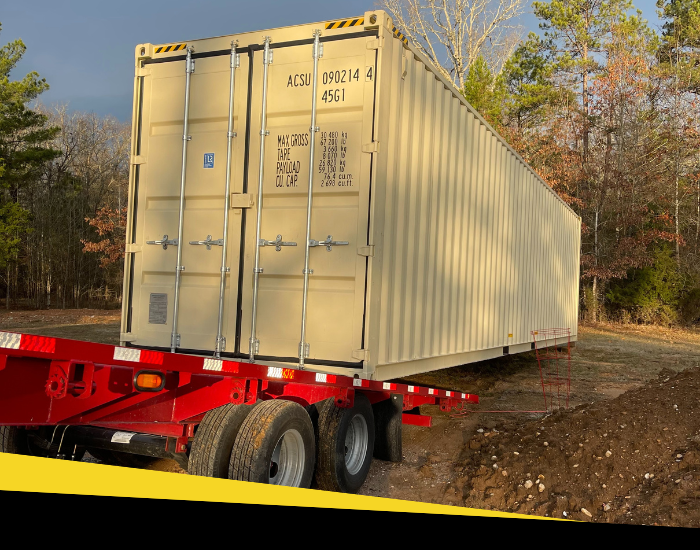 A large tan shipping container sits on a red flatbed trailer on a dirt lot surrounded by trees.
