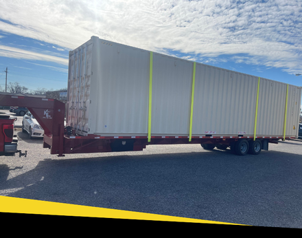 A large, light-colored shipping container secured with yellow straps on a red flatbed trailer in a parking lot.