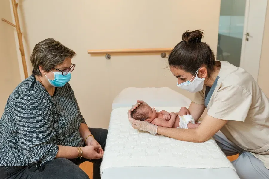 Nurse examining a newborn baby on a table; woman watches. All wear masks in a medical setting.
