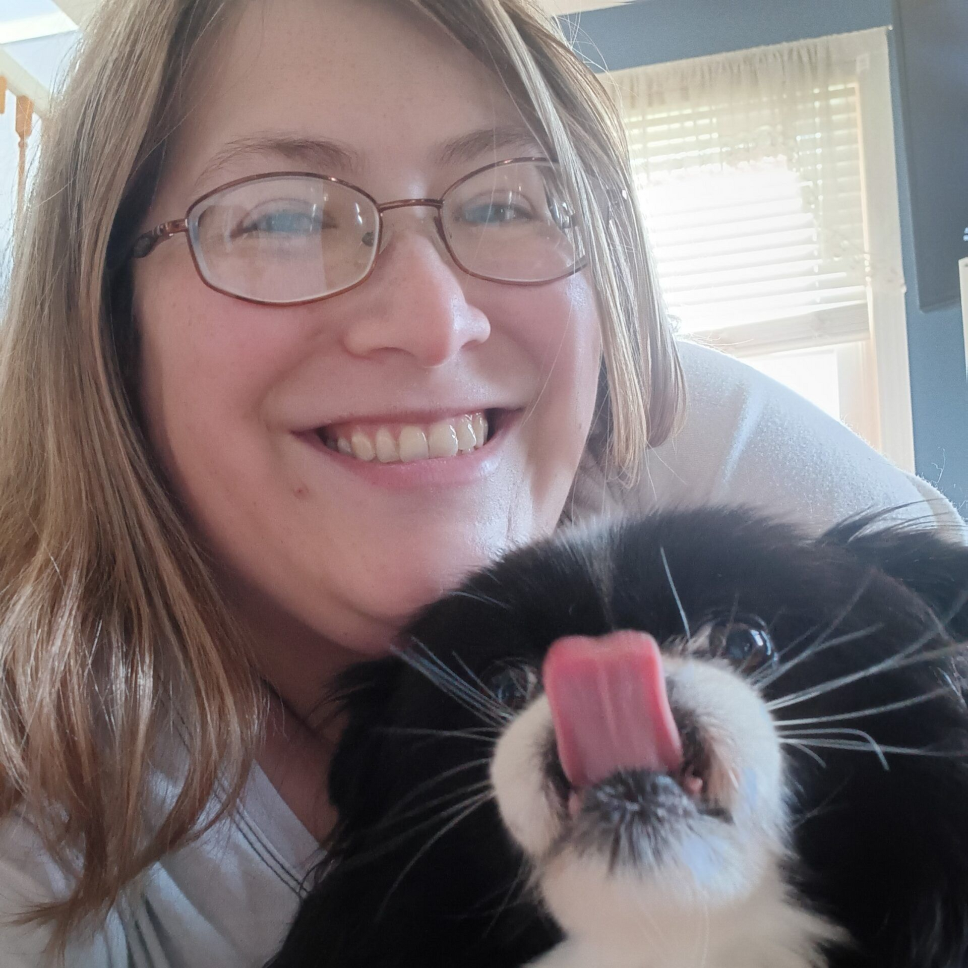 Woman in glasses smiling, holding black and white dog that licks its nose.