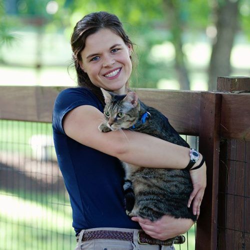 Woman in blue shirt holding a tabby cat outdoors, smiling. Brown fence and green background.