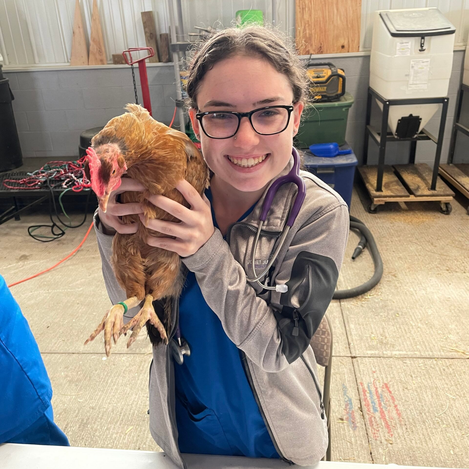 A person in scrubs smiles, holding a chicken. They are indoors.