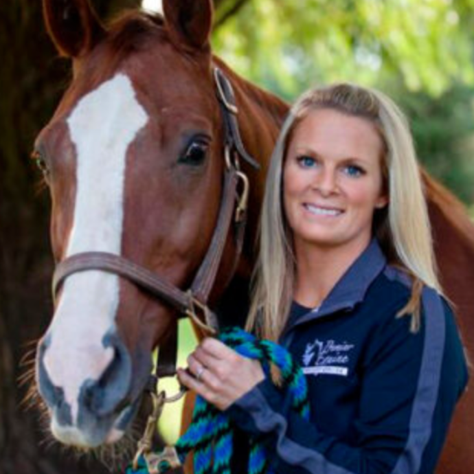 Woman with blonde hair holding a rope next to a brown horse with a white blaze in an outdoor setting.