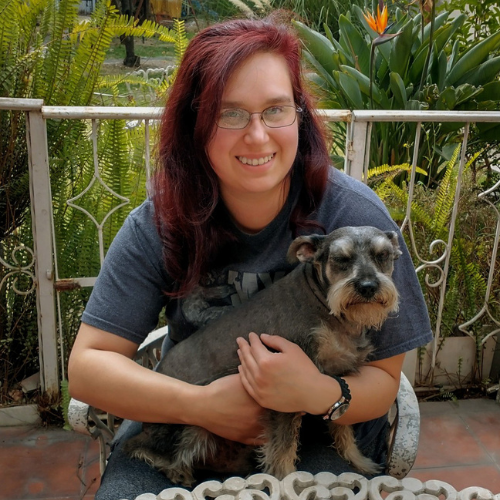 Woman with red hair, glasses, smiling, holding a gray dog. Outdoors, plants in background.
