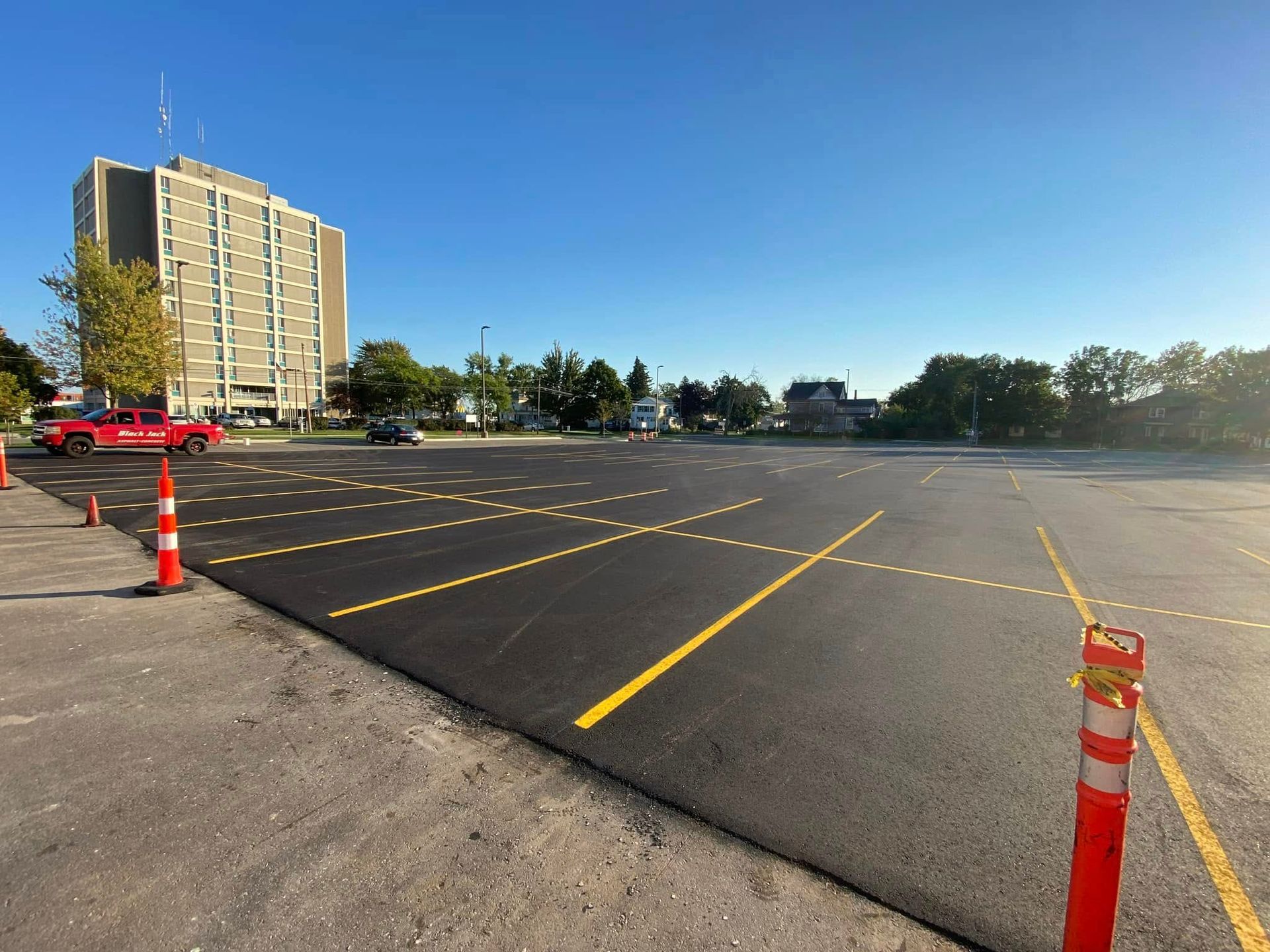 A road milling machine loads debris onto a white dump truck in a large parking lot near a multi-story building.