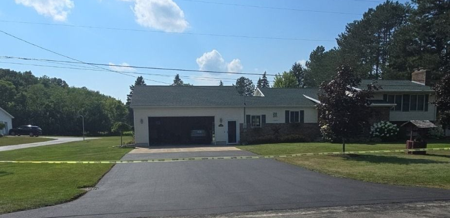 House with black asphalt driveway and garage, trees, and blue sky.