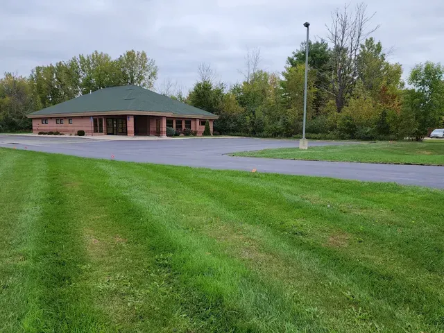 A one-story brick building with a green roof, surrounded by a large green lawn and a paved parking area.