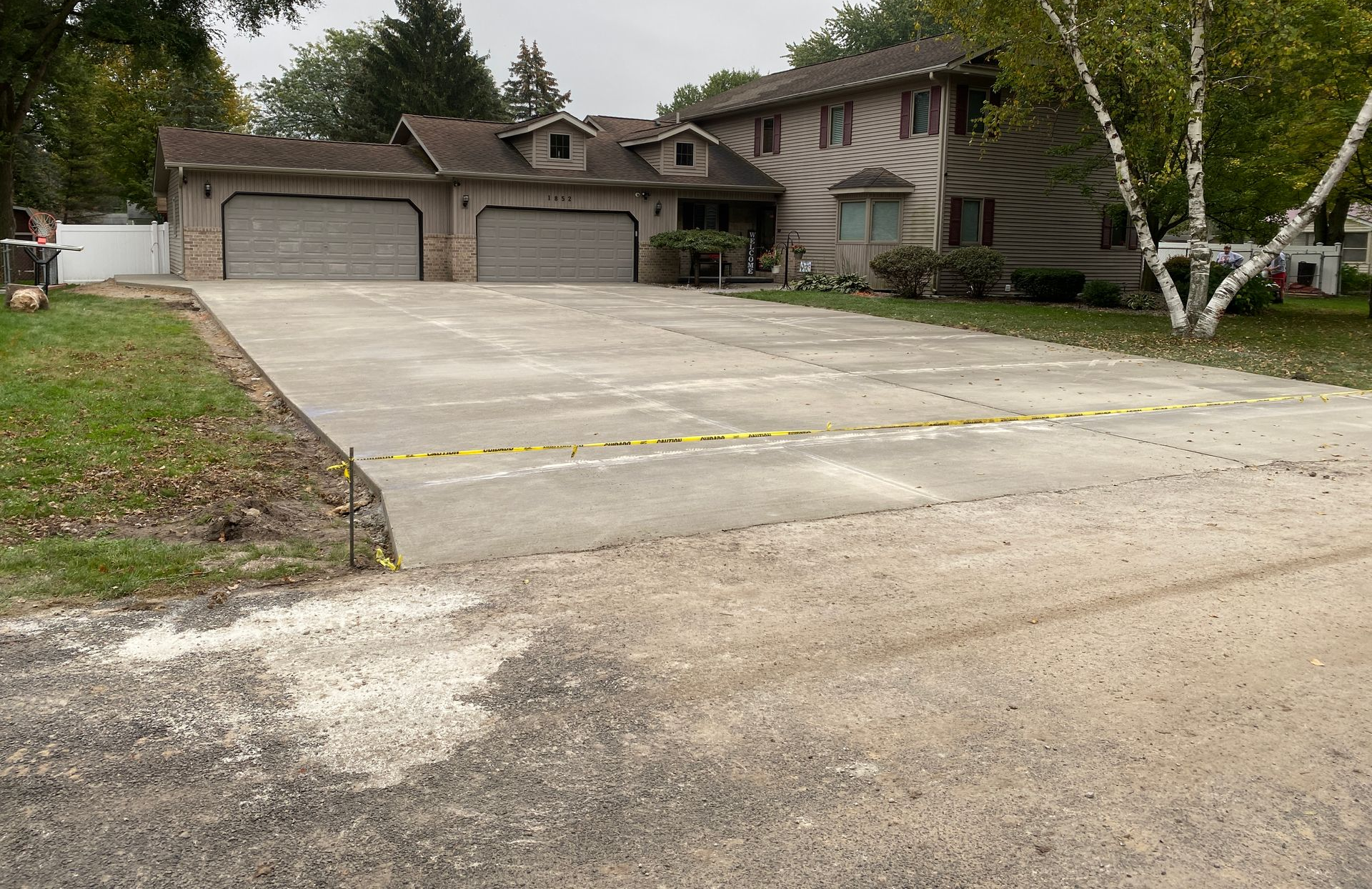 Black asphalt driveway curves through a grassy lawn with trees, under a bright, cloudy sky.