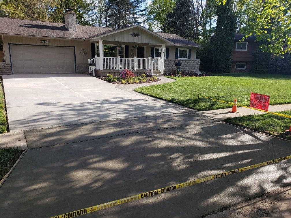 Paved driveway leading to a large house, surrounded by trees and green lawn on a sunny day.
