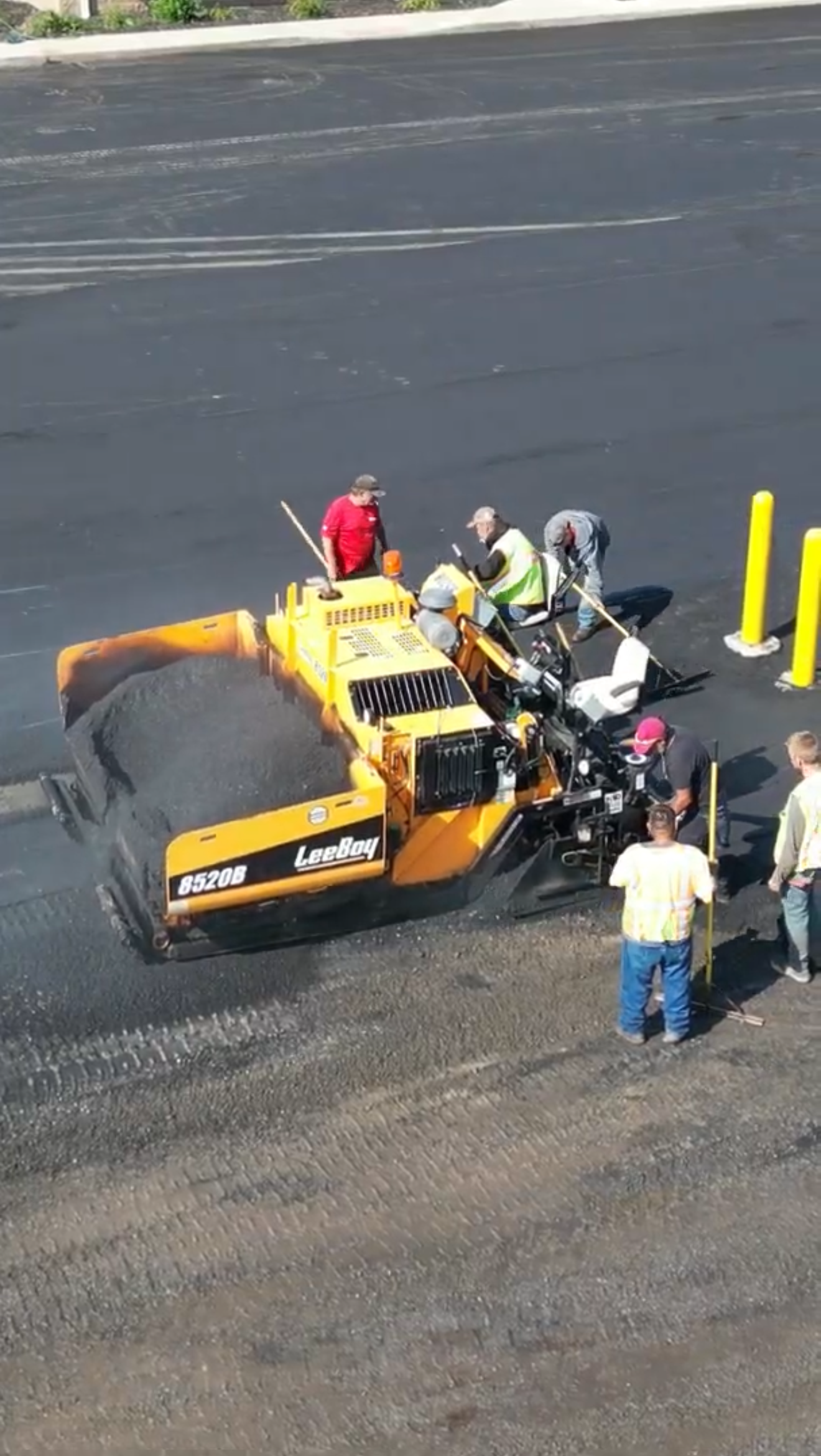 Asphalt paving construction in progress near a multi-story building.  Workers operate machinery, orange roller, paving machine.