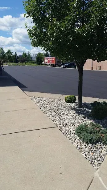 Sidewalk beside a freshly paved parking lot with a tree and landscaped stone border.