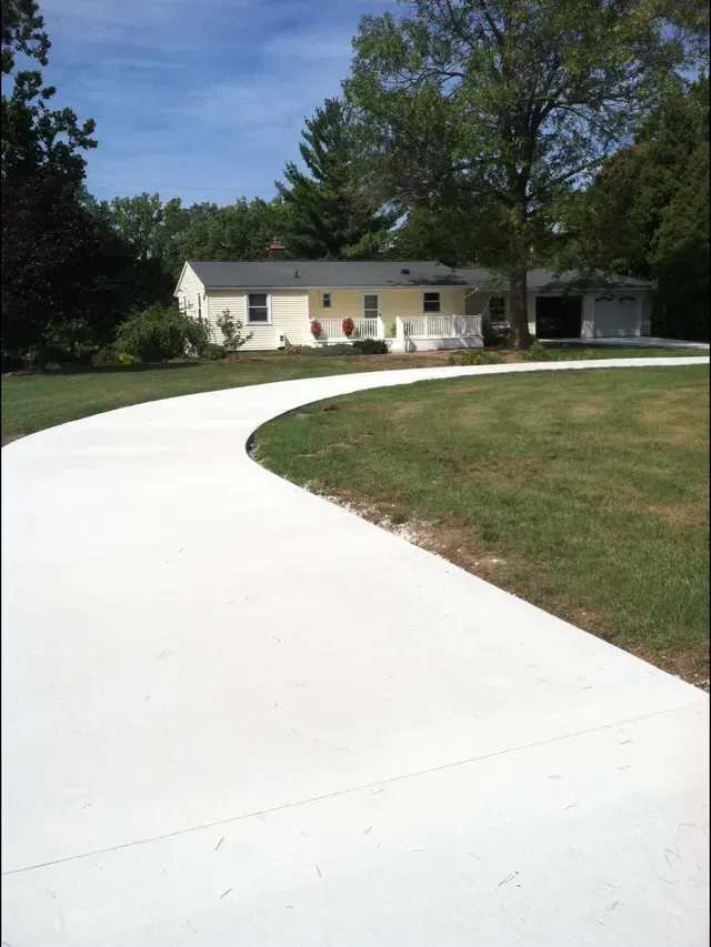 White concrete driveway leading to a light yellow house with a white porch and garage on a sunny day.