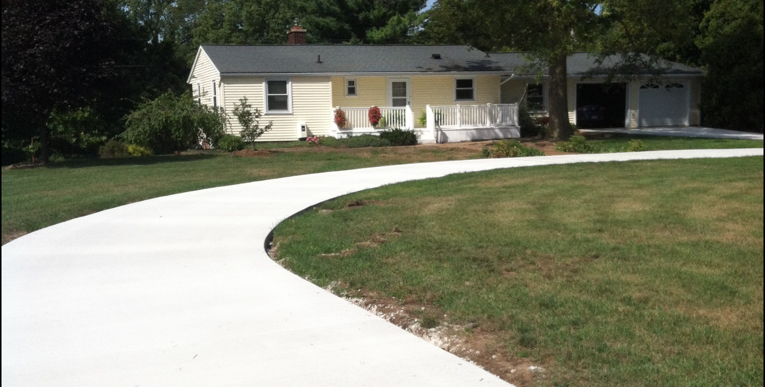 Curving concrete driveway leads to a light yellow house with white porch and attached garage.