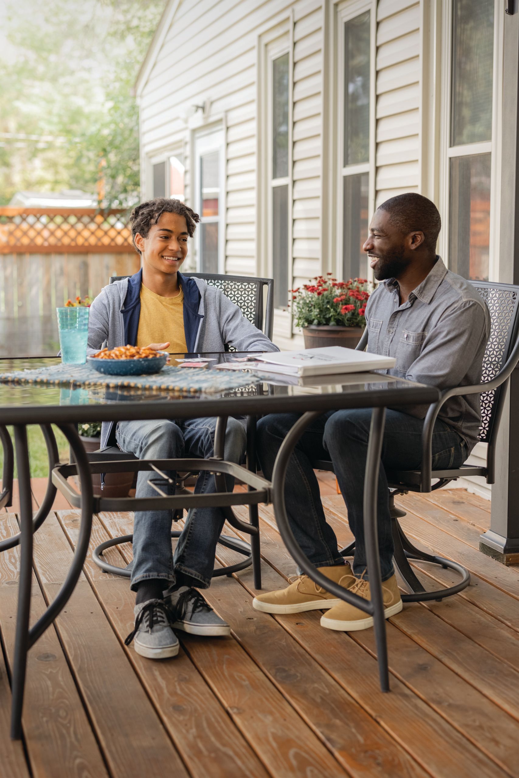 Two people on a deck; one is smiling, the other is talking. Table with food and plants.