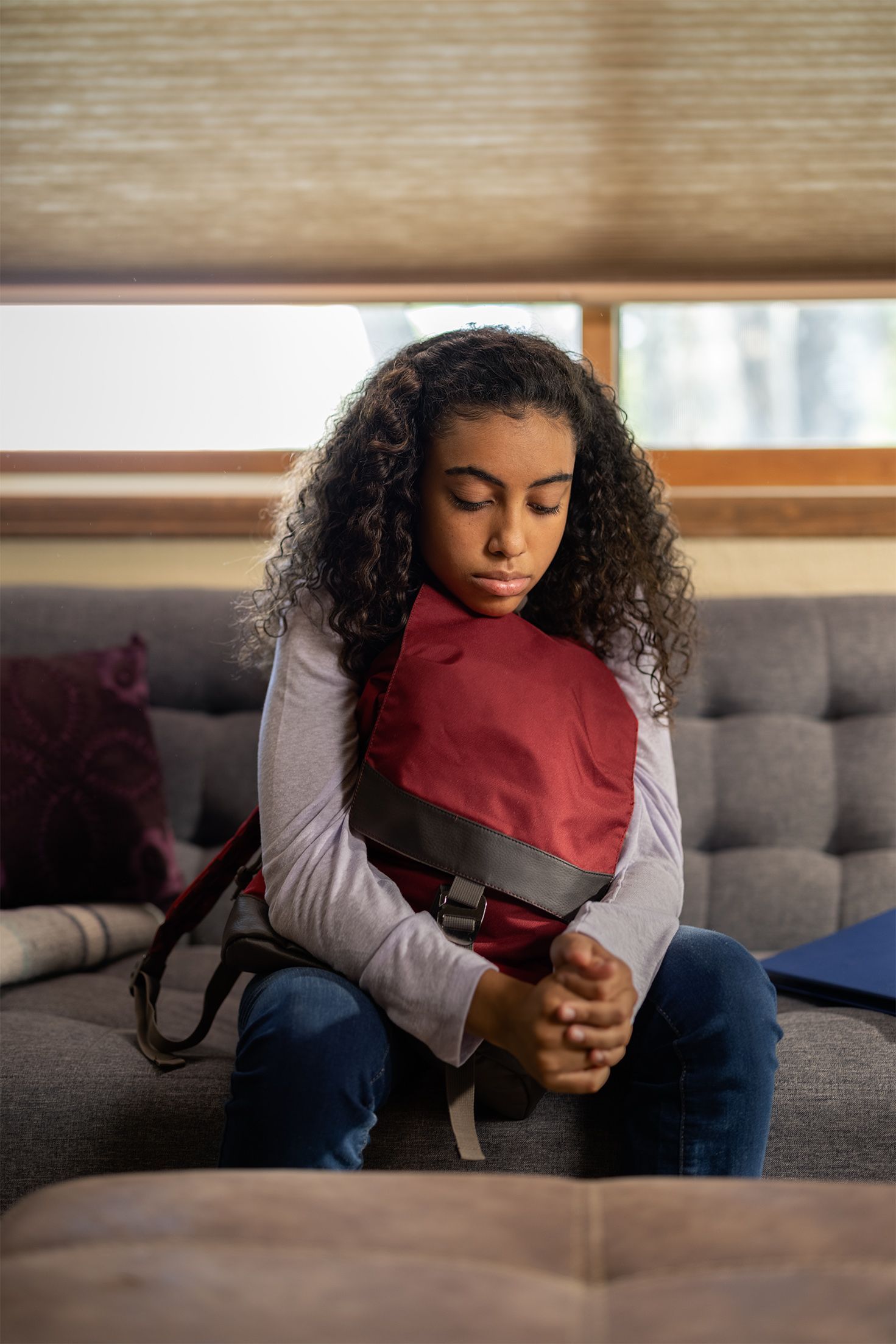 Girl with curly hair, sitting on a couch, holding a red backpack, looking down.