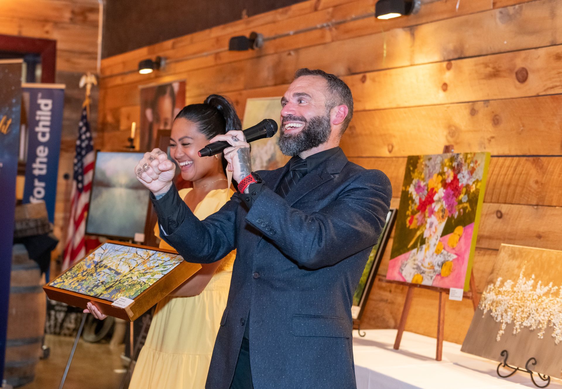 Man with microphone gestures, woman holds art at event with paintings and US flag.